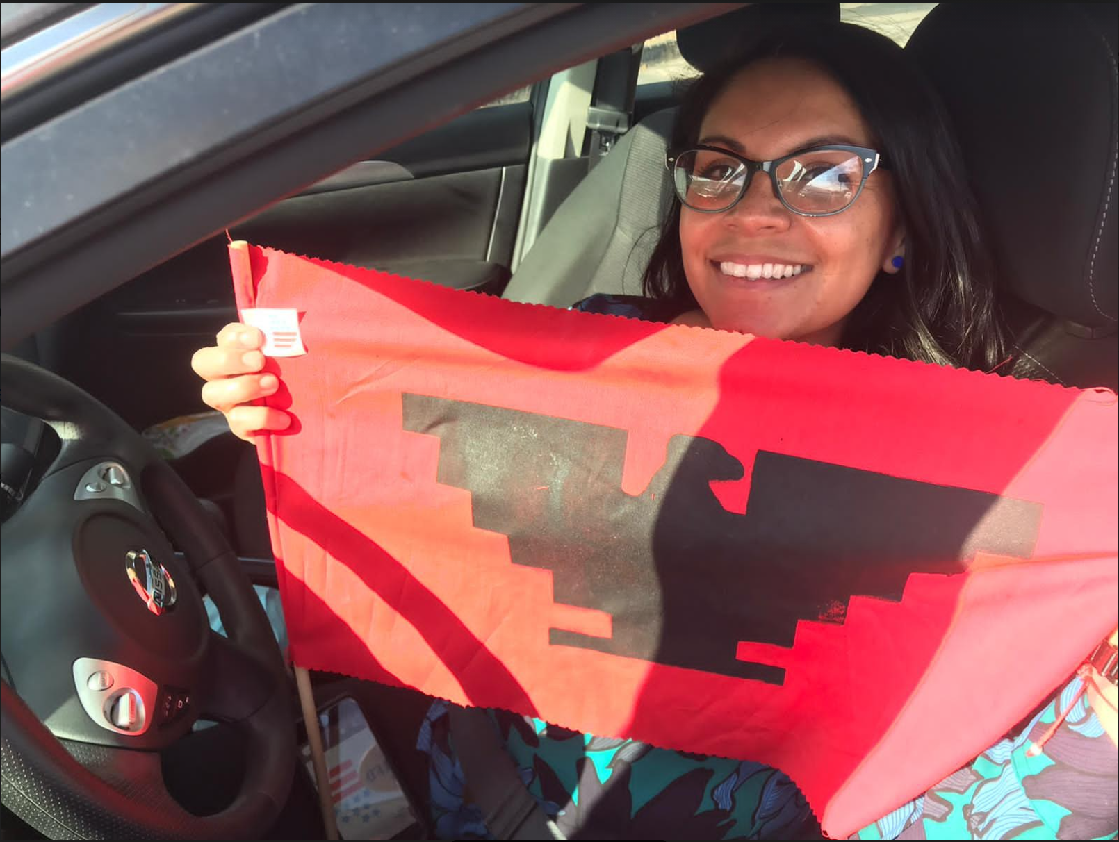 Driver sits behind the wheel smiling and holding up a red and black United Farm Worker flag emblazoned with a black aztec eagle on a red field.