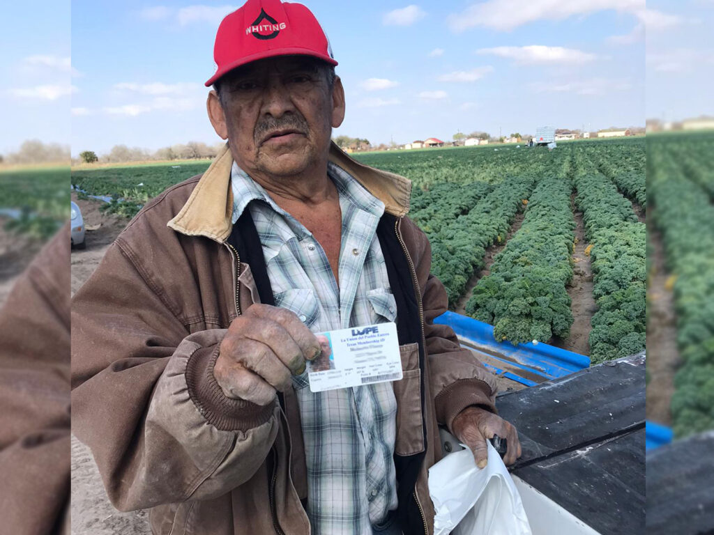 Farmworker with red cap and heavy work jacket poses in front of field furrows holding up a LUPE membership card in one hand and a bag of PPE in the other