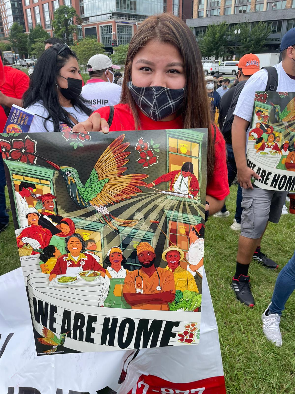 Karina Hernandez stands surrounded by other activists with signs that read "We Are Home" and show graphics of farmworkers and essential workers